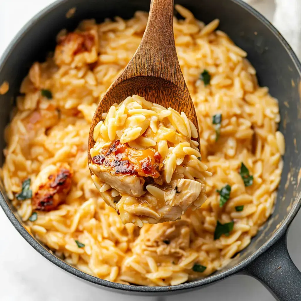 Une cuillère en bois servant des pâtes orzo crémeuses avec du poulet depuis une casserole noire.
