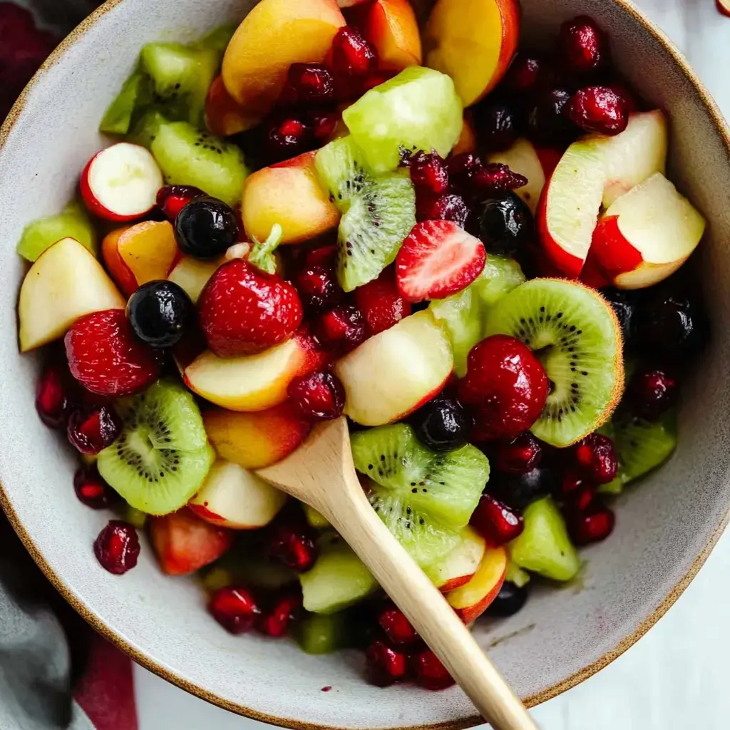 Une salade colorée avec des tranches de pommes, fraises, kiwis et melons, mélangée à des framboises et graines de grenade dans un bol avec une cuillère en bois.