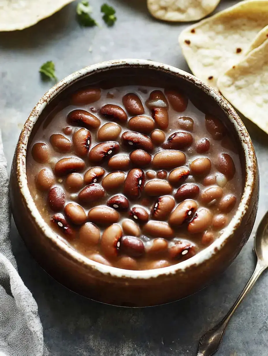 Un bol de haricots cuits dans un bouillon, des tortillas fraîches et de la coriandre par-dessus, sur une table rustique.