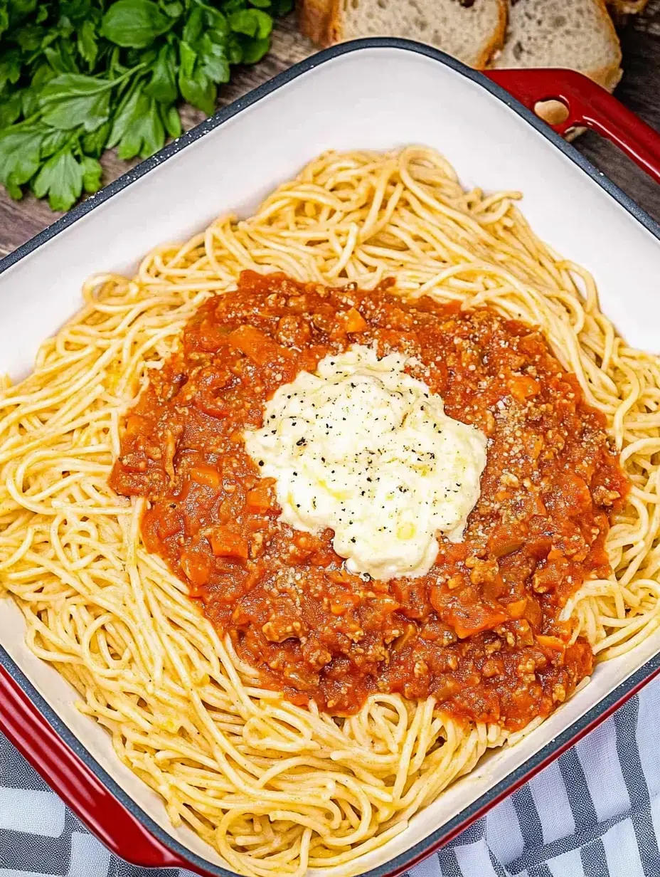 Zoom sur un gratin de spaghetti nappé de sauce viande et d'une couche crémeuse, présenté dans un plat carré avec du persil frais et une baguette en arrière-plan.