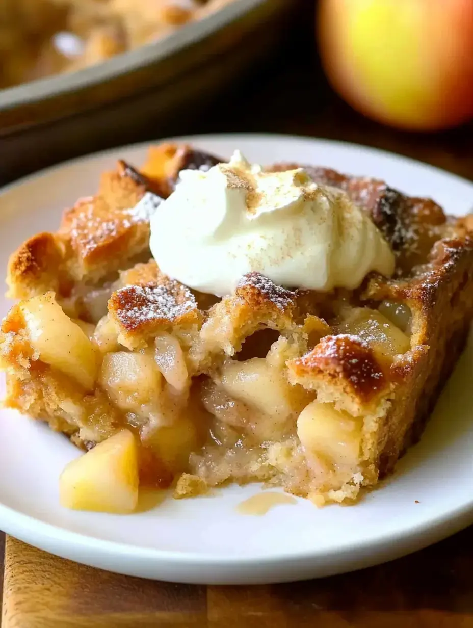 Une part de dessert pomme-brioche sur une assiette, surmontée de chantilly et saupoudrée de sucre glace, avec une pomme à côté.