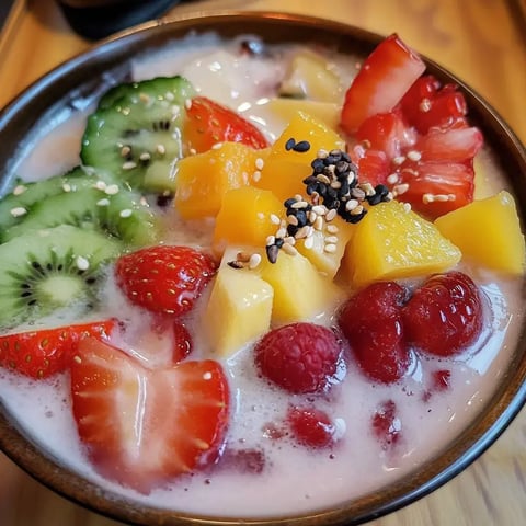 Un bol de fruits frais variés, comme des fraises, du kiwi, de la mangue et des framboises, servis dans une base crémeuse façon yaourt avec des graines de sésame parsemées au-dessus.