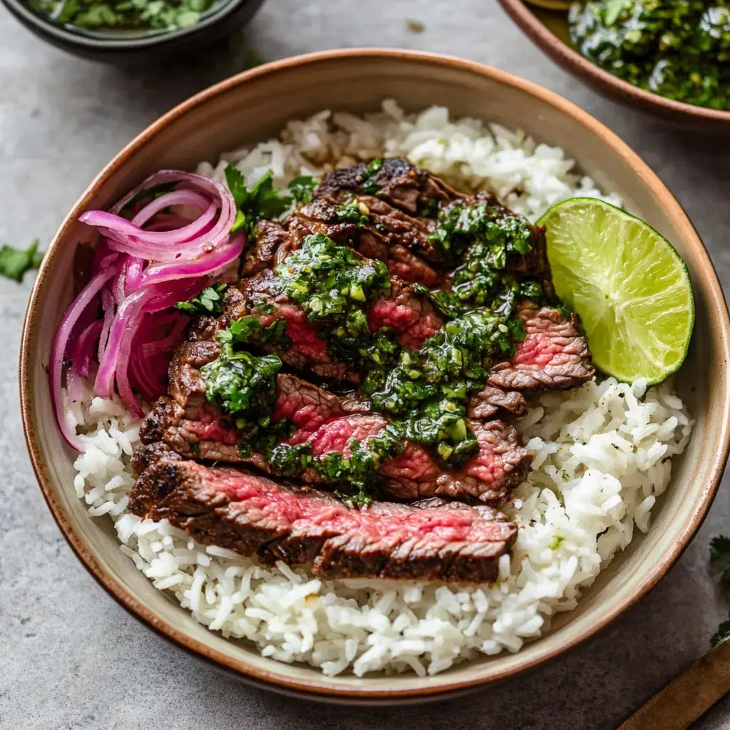 Un bol de riz garni de tranches de bavette, sauce chimichurri, oignons rouges marinés et une rondelle de citron vert.