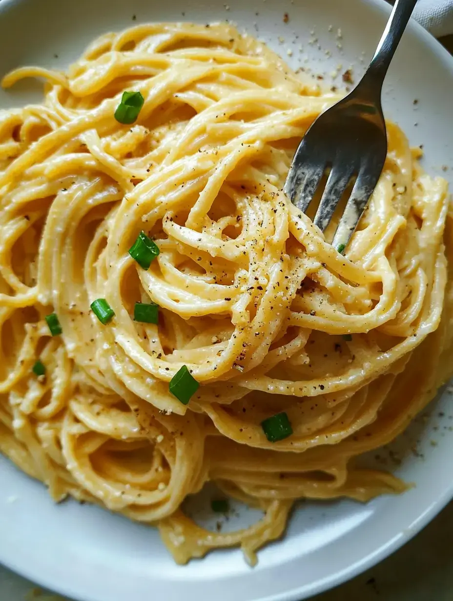Un gros plan de pâtes crémeuses garnies d'oignons verts et de poivre noir dans une assiette blanche, avec une fourchette posée dans les nouilles.